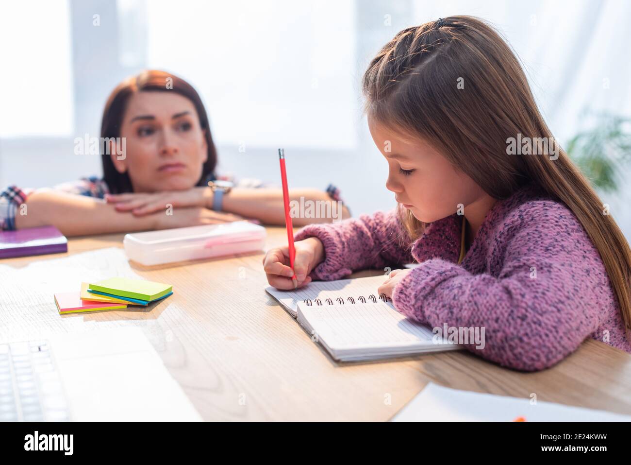 Upset mother looking at daughter writing in notebook at desk on blurred background Stock Photo
