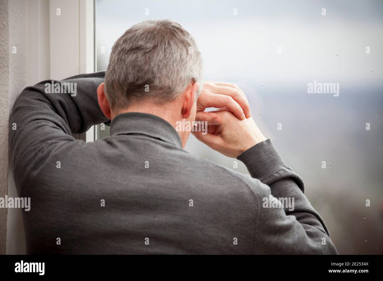 Sad mature man leaning on a window with raindrops - focus on the hands and the window Stock Photo