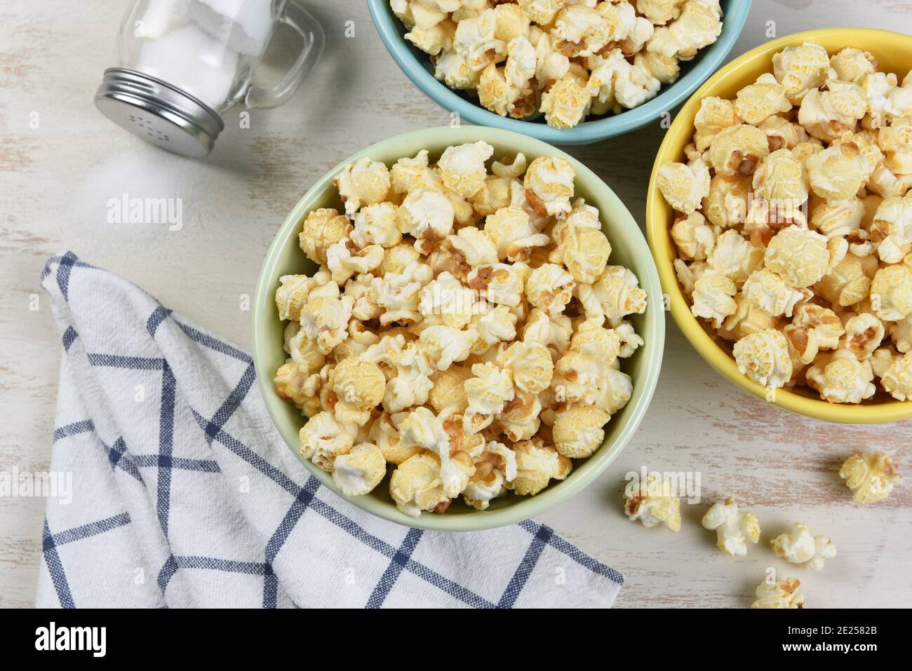 High angle shot of three different colored bowls of fresh popped popcorn with towl and salt shaker with salt spilling out. Stock Photo