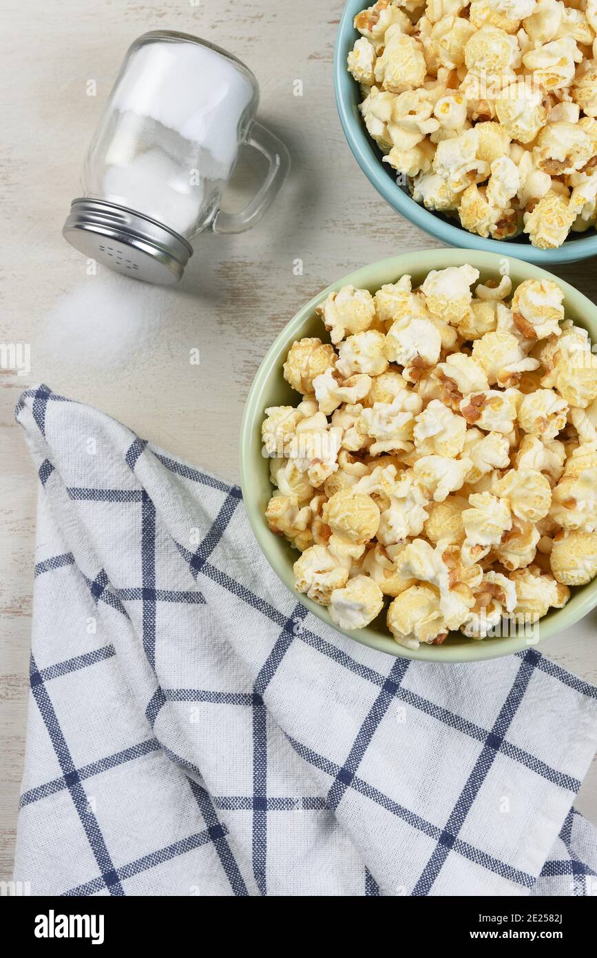 High angle shot of two different colored bowls of fresh popped popcorn with towl and salt shaker with salt spilling out. Vertical format. Stock Photo