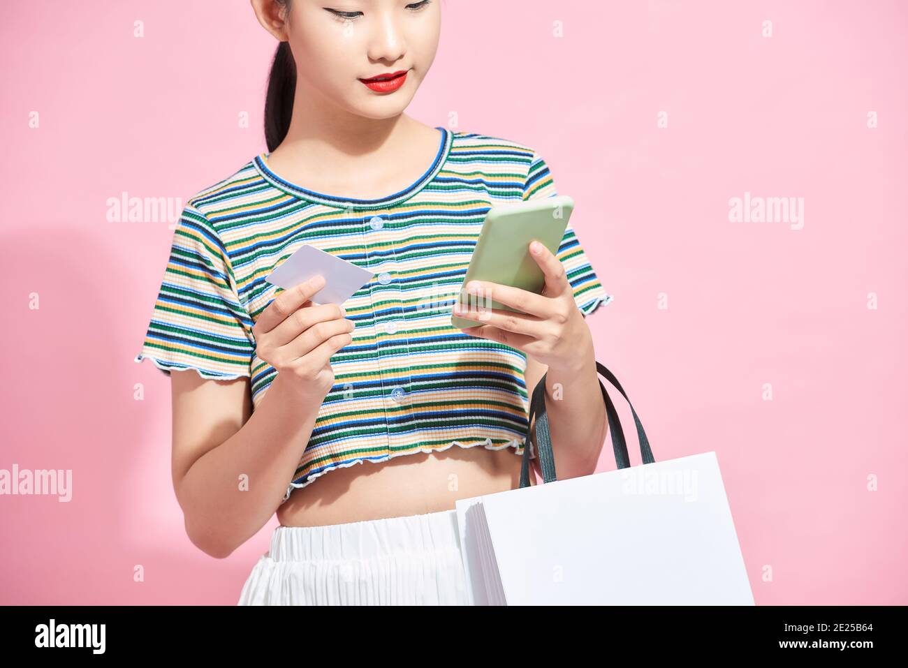 Young smiling beautiful Asian woman making online payment through mobile phone via credit card while carrying colorful shopping bags Stock Photo