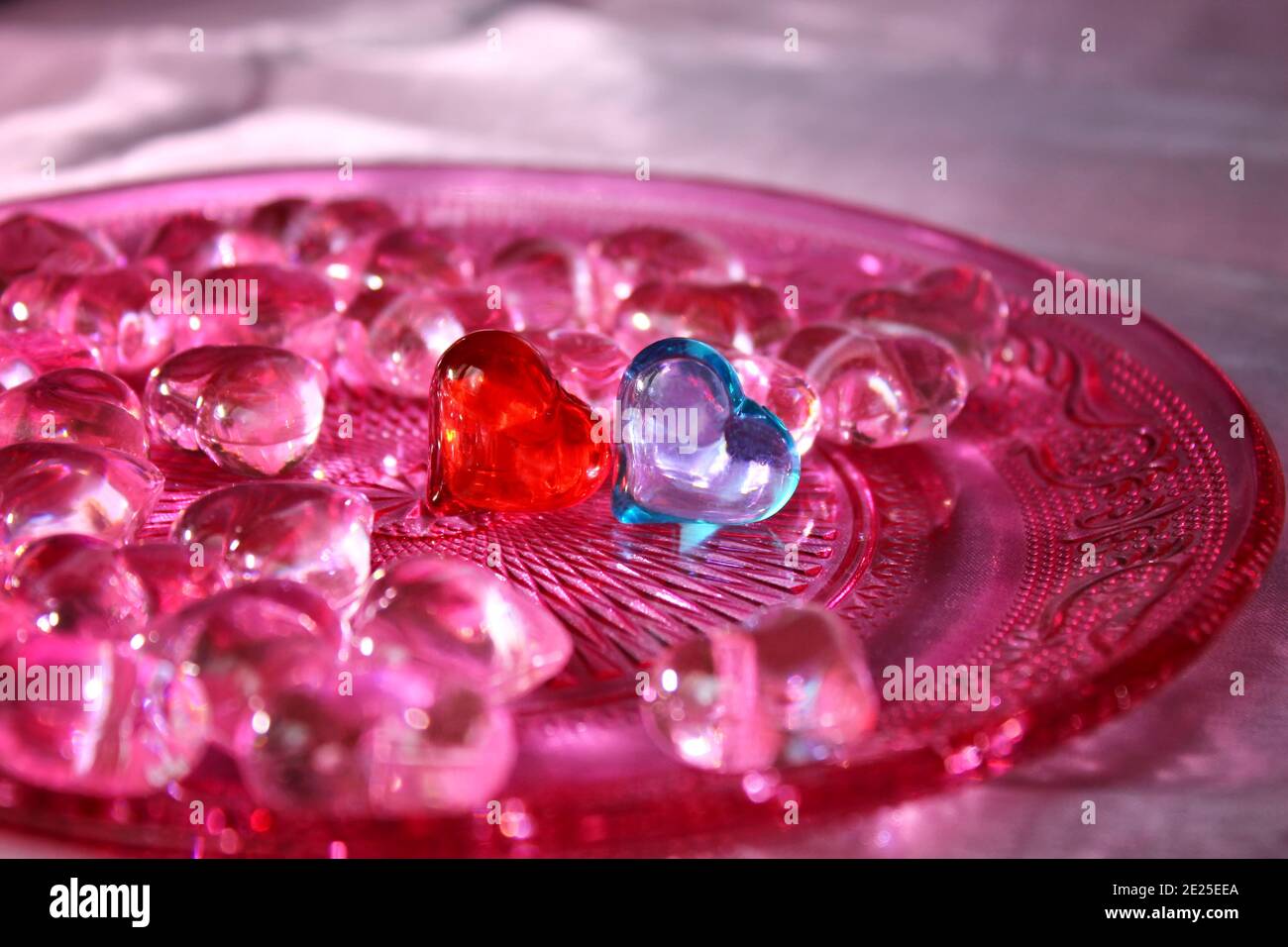 A red and a blue glass heart surrounded by pink hearts on a pink glass plate with ornament pattern. Stock Photo