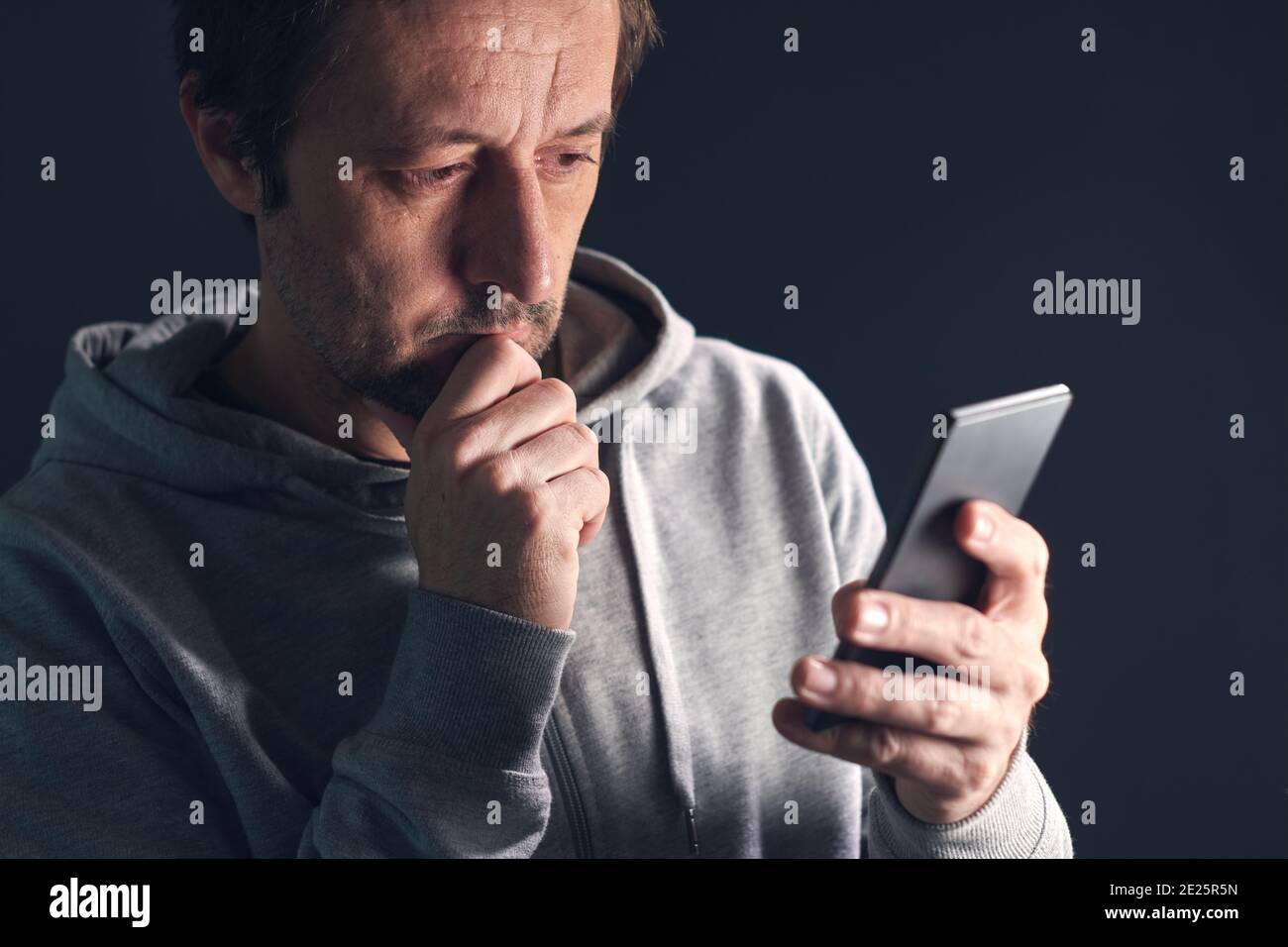Worried casual mid-adult man looking at the screen of his smartphone with one hand on the chin Stock Photo