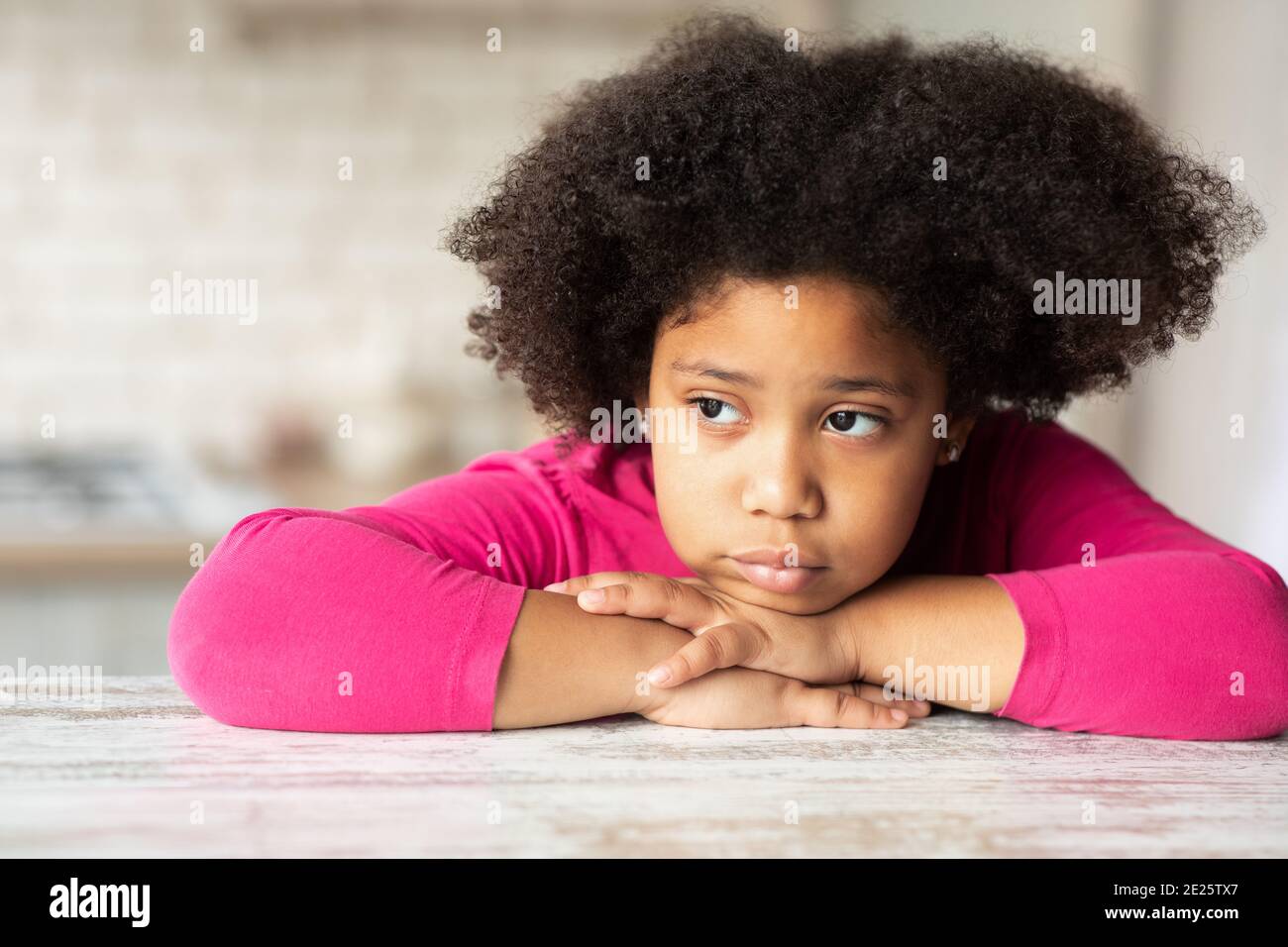 Portrait of cute sad little black girl sitting at table in kitchen. Stock Photo