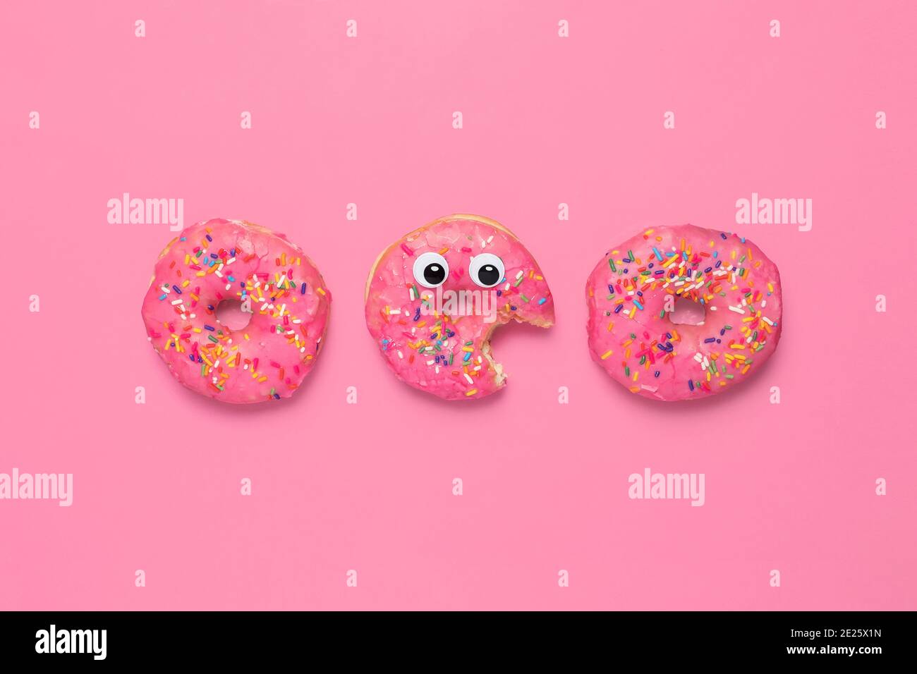 Three pink donuts doughnuts on a pink background.  One has googly eyes and a bite taken out of it.  Copy space and room for text Stock Photo