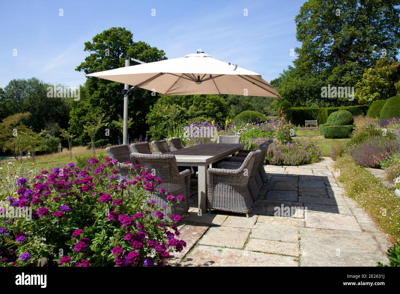 Garden terrace with large dining table and ten chairs and a large rectangular umbrella parasol with flowers, hedging and trees Stock Photo