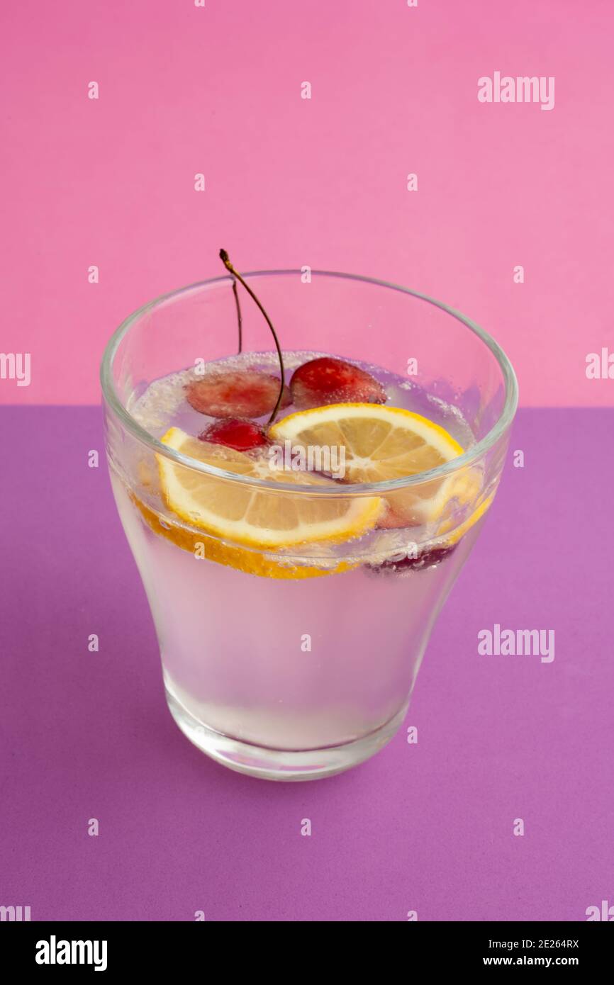 a glass of sparkling water cocktail with sliced lemon, cherry and berry. non-alcoholic drink, beverage. purple textured backdrop and pink background. Stock Photo