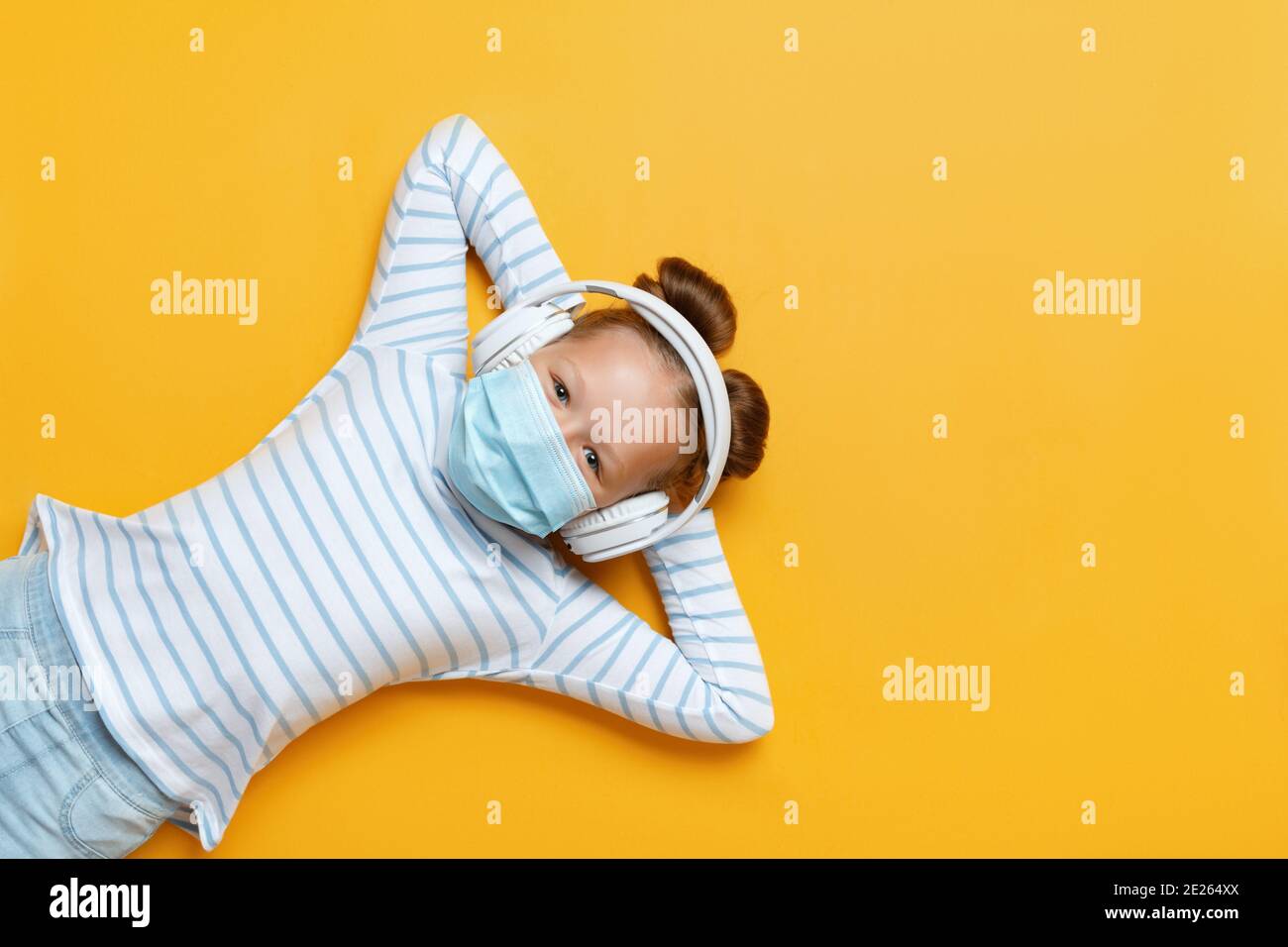 Cheerful cute little girl listening to modern technology headphones on yellow background. The child lies in a protective medical mask, hands behind hi Stock Photo