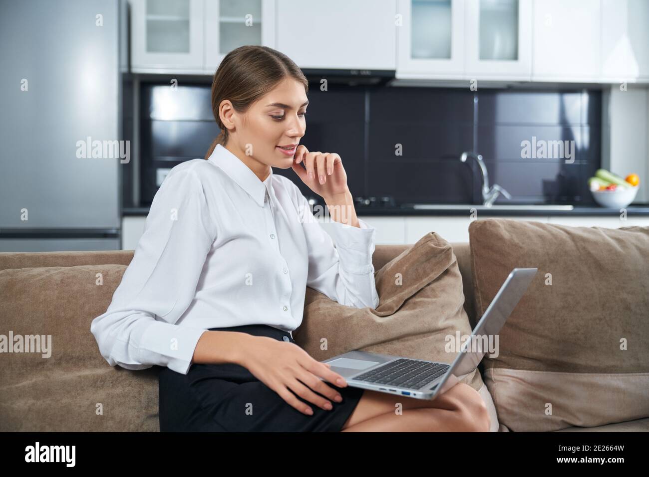 Smiling young lady relaxing on cozy sofa with portable laptop on knees. Attractive woman in white shirt looking at computer screen at home. Stock Photo