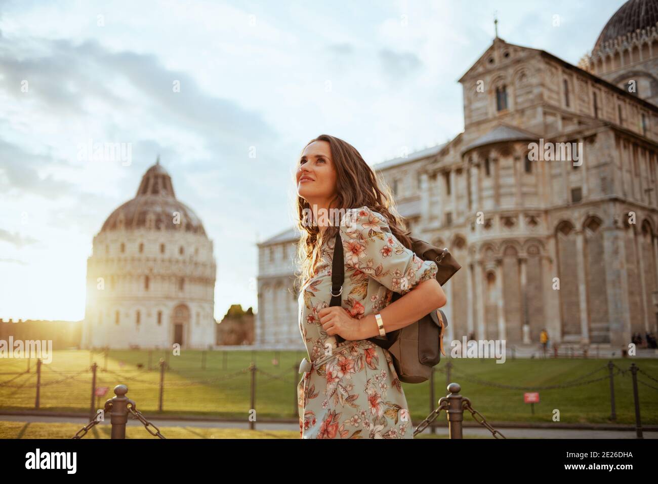 smiling trendy traveller woman in floral dress with backpack enjoying promenade in piazza dei miracoli in Pisa, Italy. Stock Photo