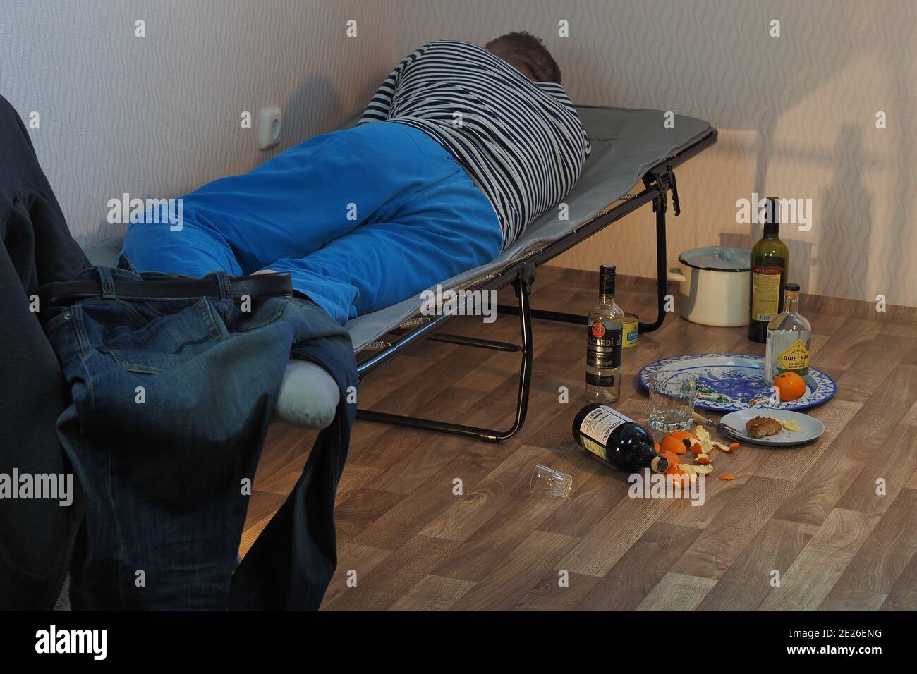 A drunk man lies on a cot and an empty bottle of alcohol is scattered nearby. Stock Photo
