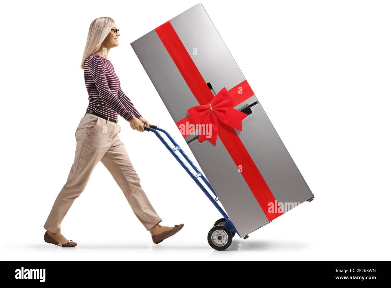 Full length profile shot of a young woman pushing a fridge with a red bow on a hand-truck isolated on white background Stock Photo