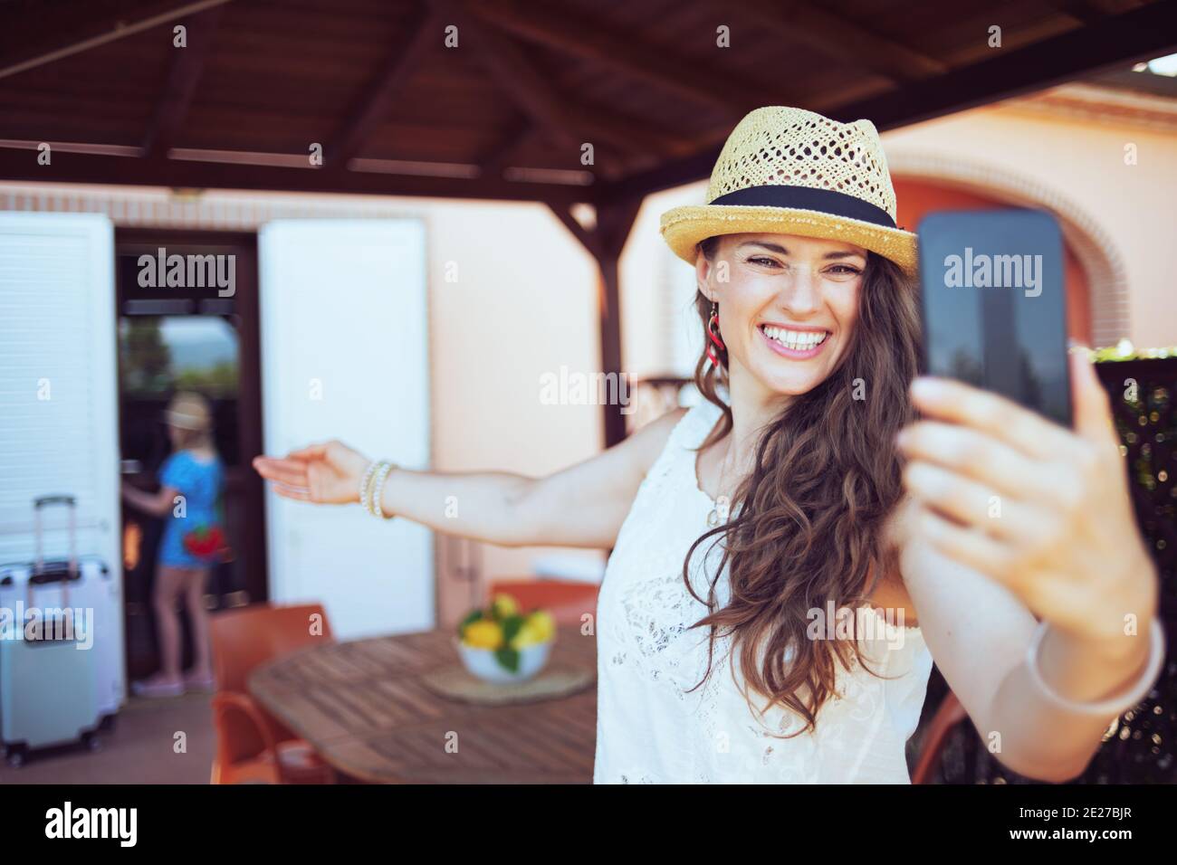 happy modern mother and child travellers in hats with wheel bags having virtual meeting on a smartphone in the terrace of guest house hotel. Stock Photo