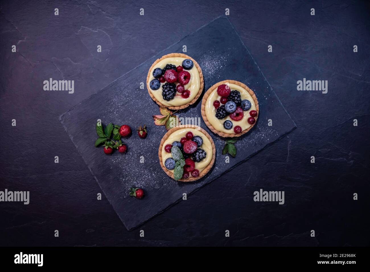 Top down view of three pie cakes with berry fruits and vanilla cream on a slate stone serving platter surrounded by strawberries. Stock Photo