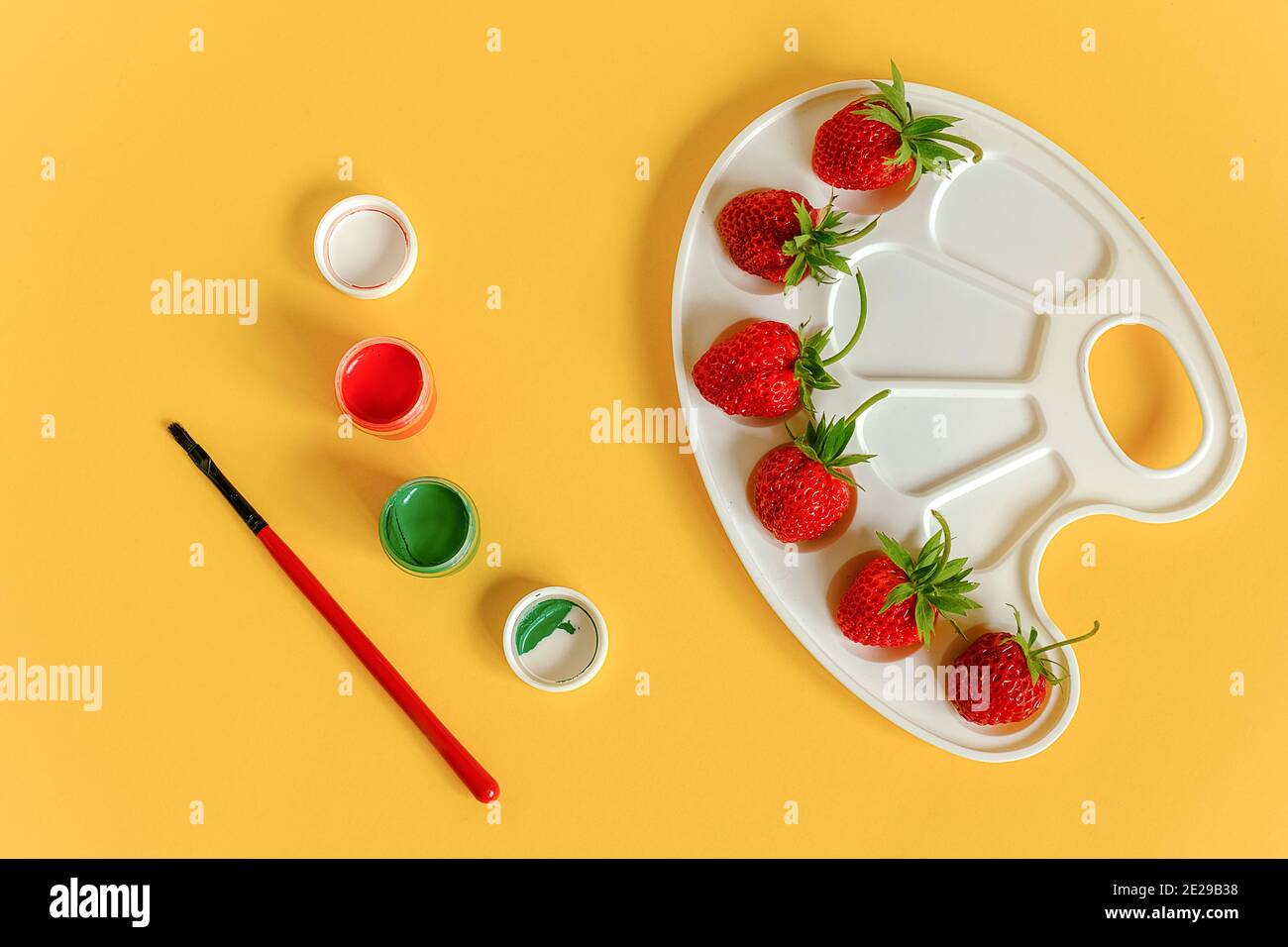 Red ripe strawberries on an artistic palette, brush and gouache on yellow background with copy space. Creative concept summer colors paint. Top view F Stock Photo
