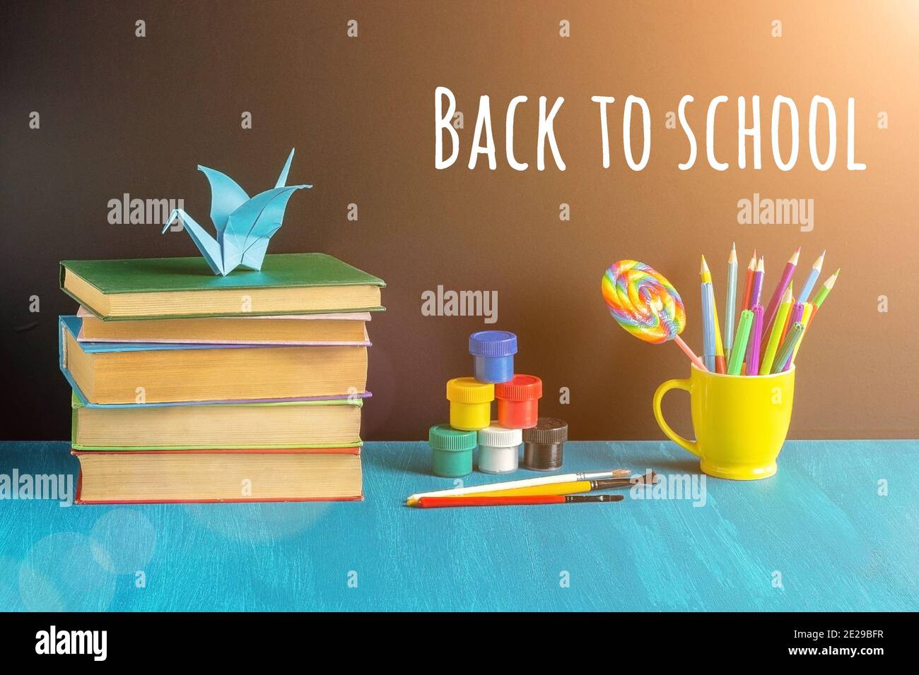 Back to school text on black chalkboard, books, stationery in yellow mug, paint gouache and origami crane on blue table. Concept Education and Pupils Stock Photo