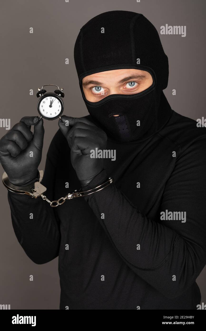 Picture of young man with black mask and outfit suspect of a robbery, wearing handcuffs in front of grey background Stock Photo