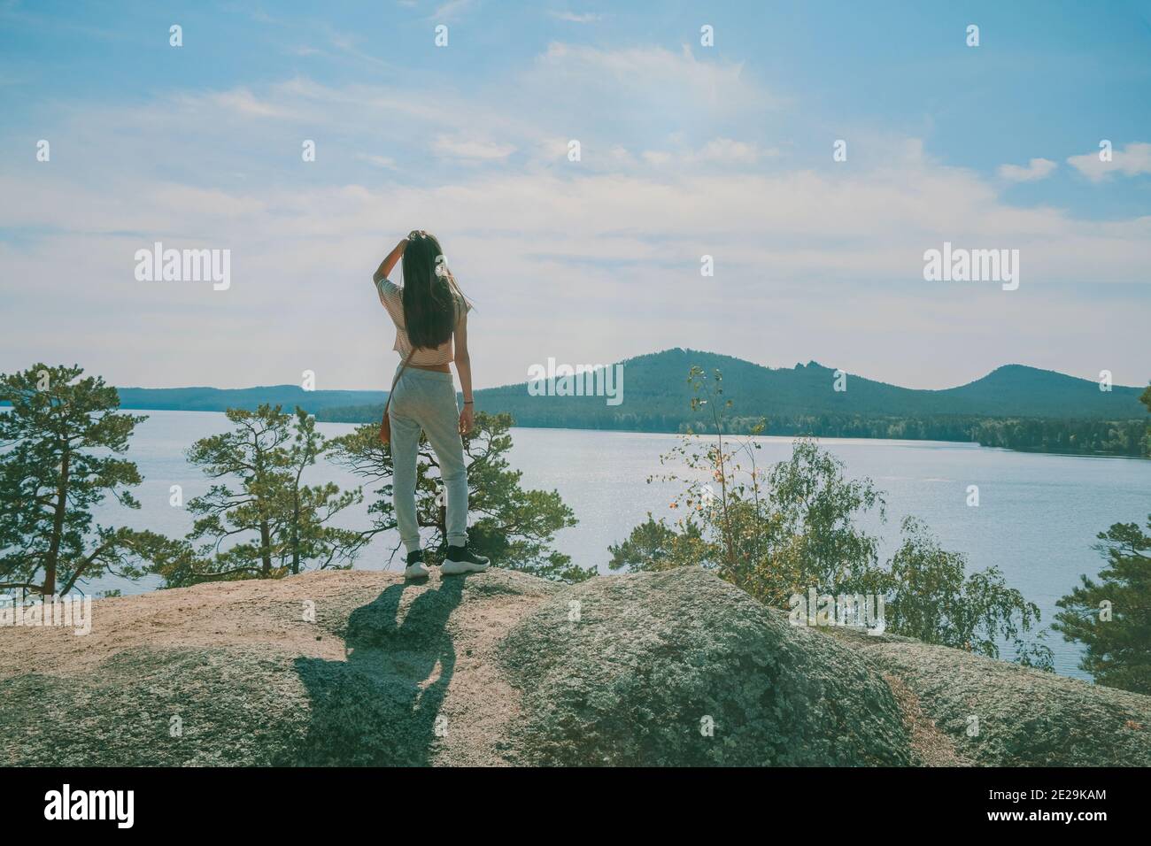 Young beautiful girl in sportswear stands on the top of mountain and looks at the lake and mountains against the blue sky. Outdoor activity or hiking Stock Photo