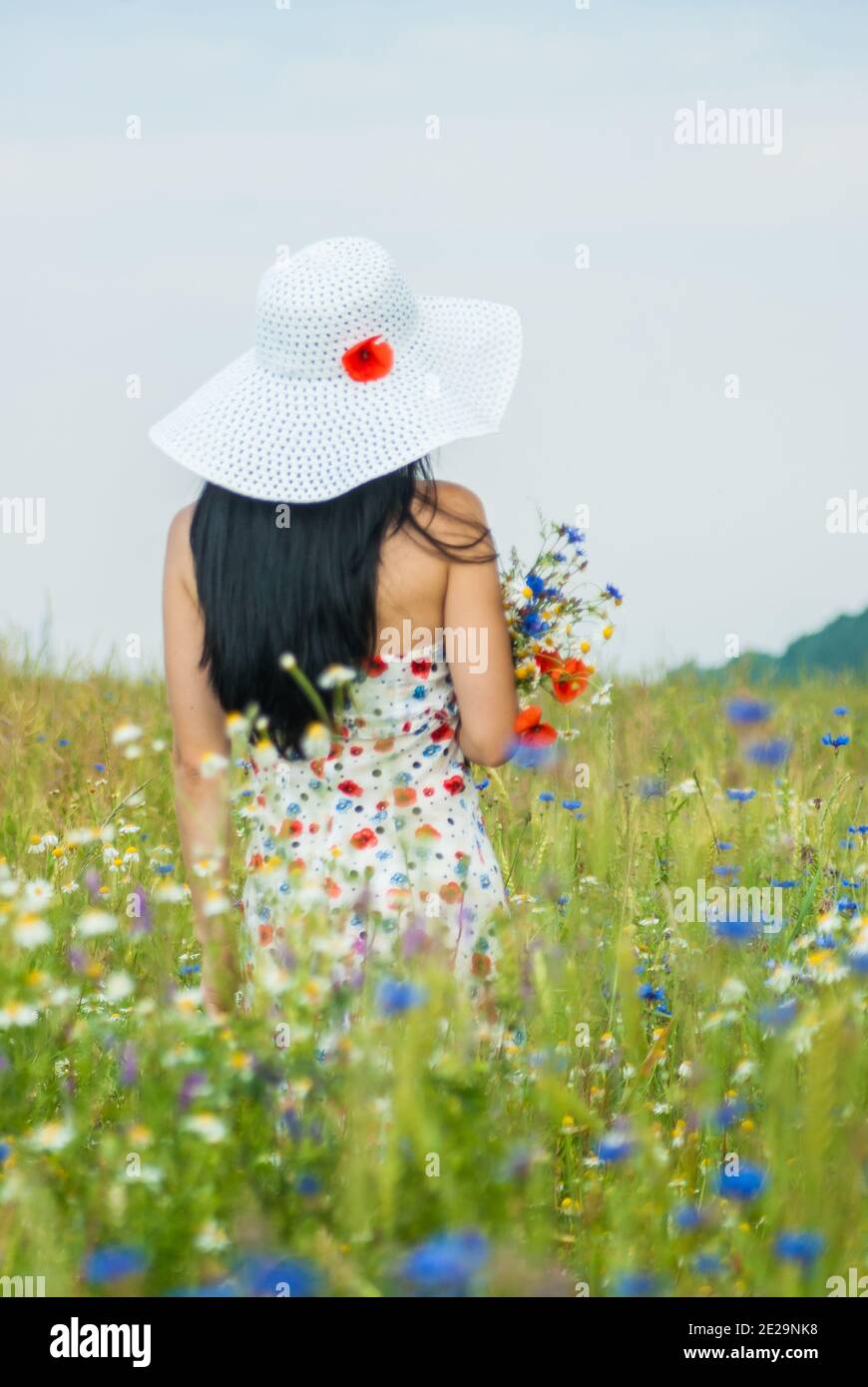 Girl in a dress and a white hat stands in a field with a bouquet of wildflowers Stock Photo