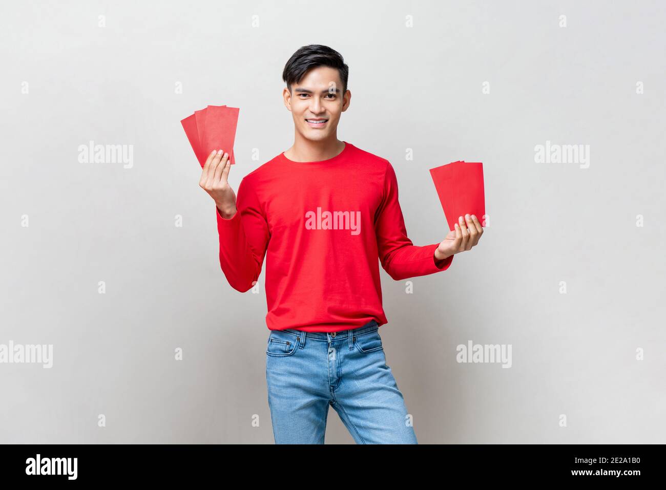 Smiling handsome Asian man holding red envelopes or Ang Pao in studio isolated gray background for Chinese new year concepts Stock Photo