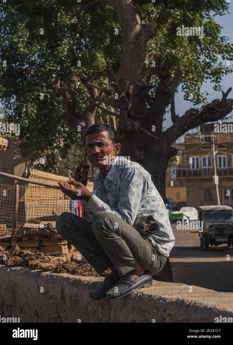 Jaisalmer, Rajasthan / India - January 8 2020 : man sitting on a wall does multitask, talking to mobile phone smoking cigarette looking at the photogr Stock Photo