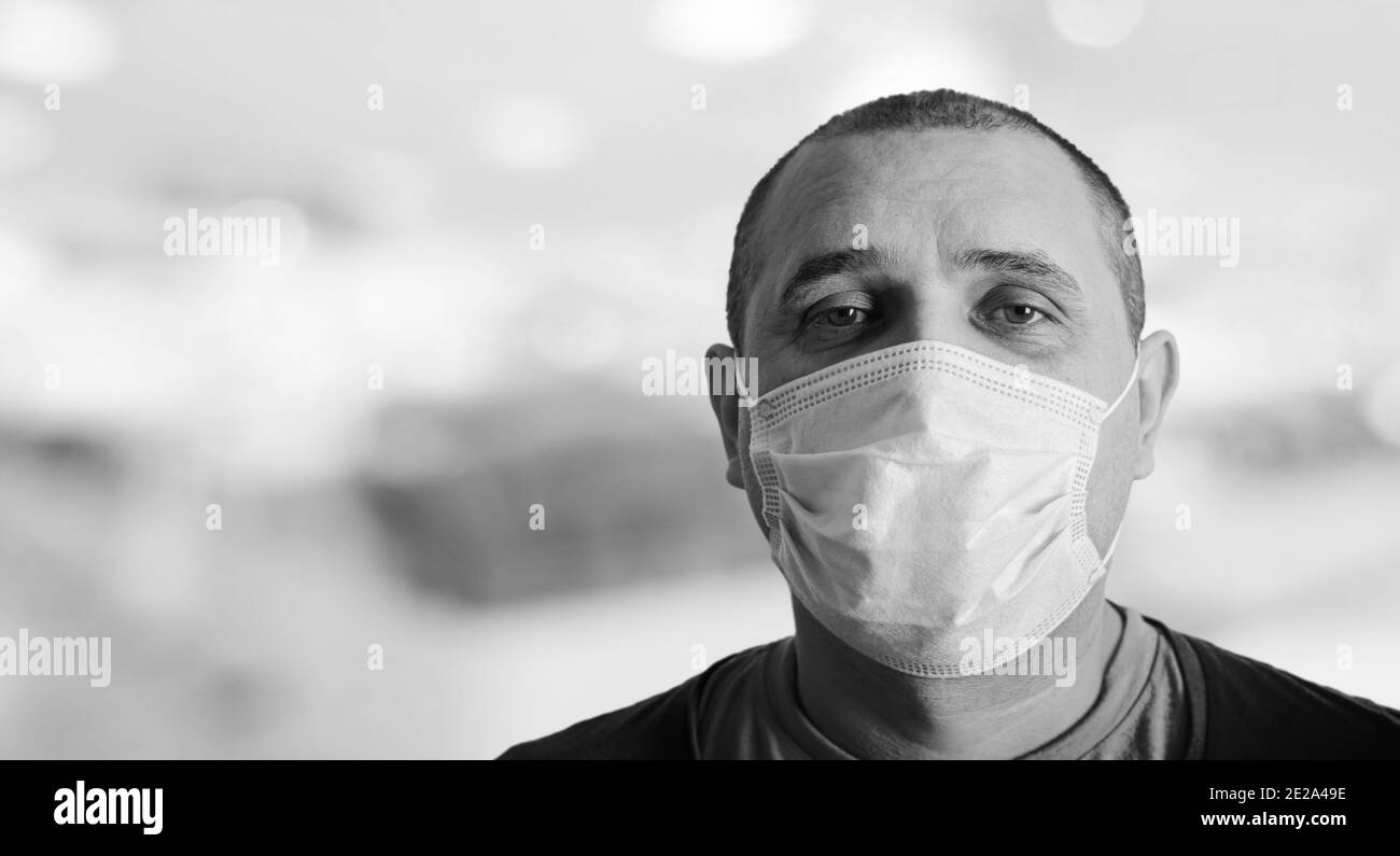 Close-up portrait of serious-looking man wearing disposable protective medical face mask on blurred hospital background. Photo in black and white colo Stock Photo