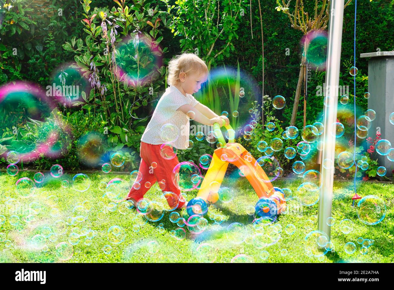Child pushing wheeled toy in lush green backyard on sunny day. Soap bubbles in foreground. Profile view. Full length. Stock Photo