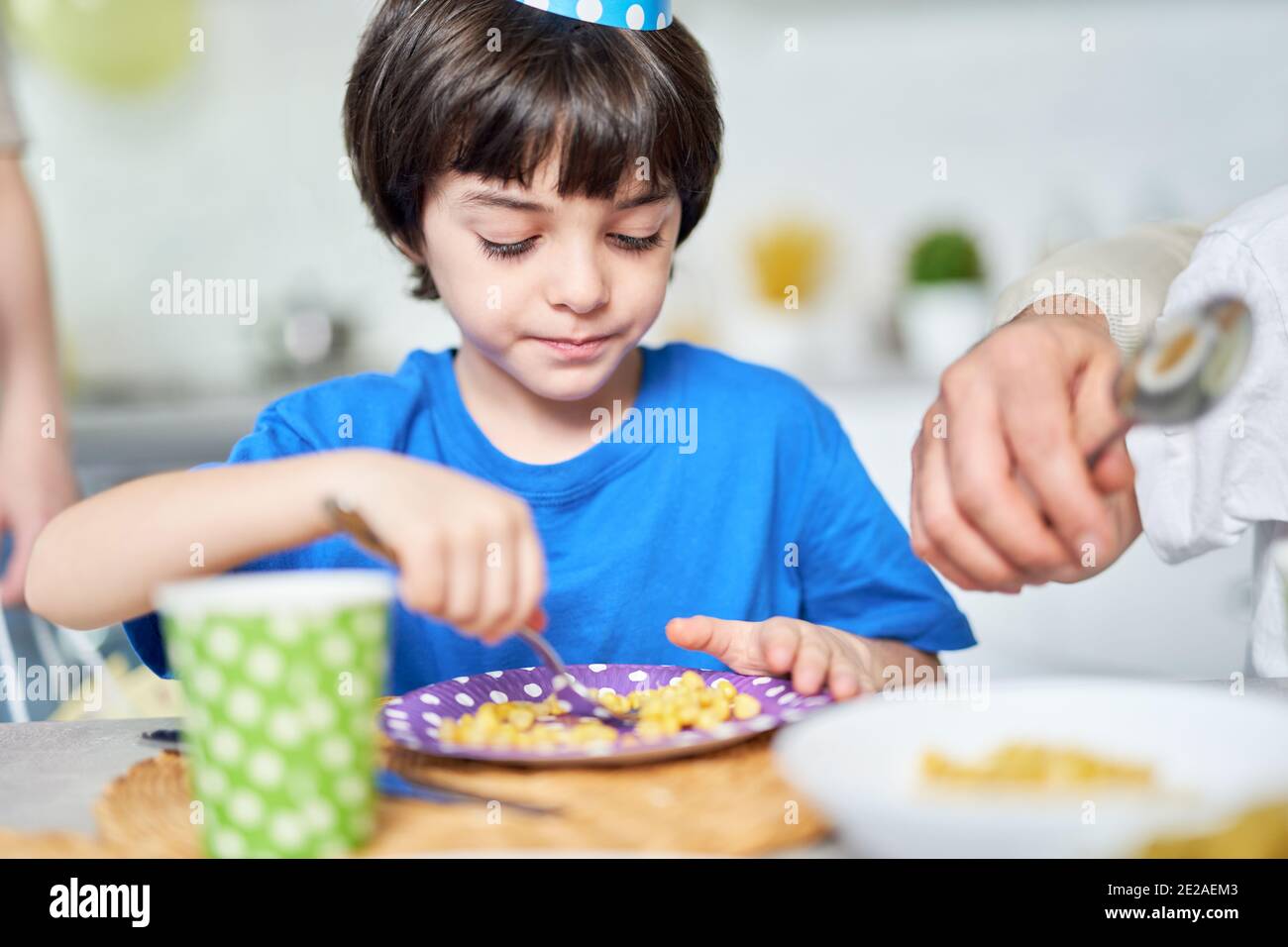 Adorable little latin american boy in birtday cap eating while celebrating birthday together with his family at home. Kids, celebration concept Stock Photo