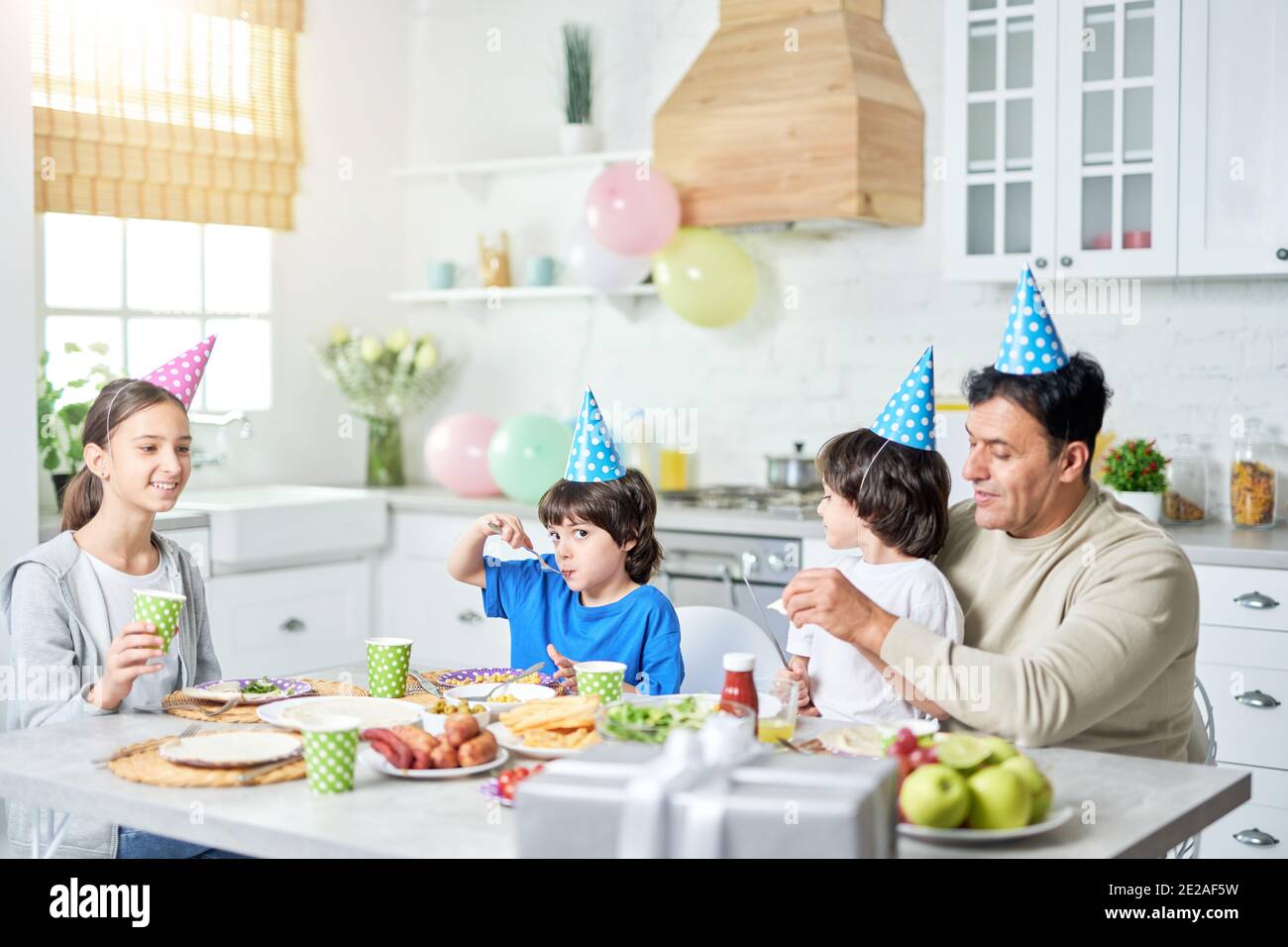Loving middle aged latin father with children wearing birthday caps on heads and having a dinner while celebrating birthday together at home. Fatherhood, happy family concept Stock Photo
