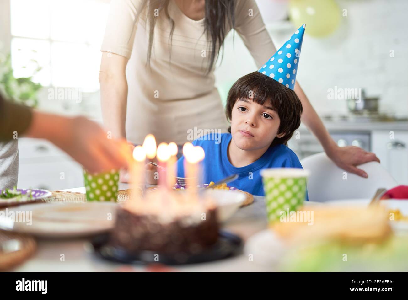 Cute little latin american boy in birtday cap looking at birthday cake while celebrating birthday with his family at home. Kids, celebration concept Stock Photo