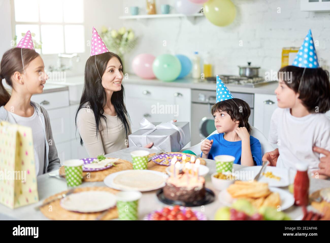 Cheerful middle aged latin mother holding gift box, talking to her lovely children while having dinner, celebrating birthday together at home. Parenthood, celebration concept. Selective focus Stock Photo