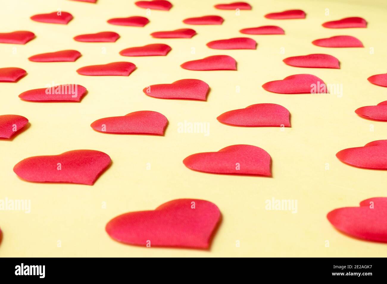 A lot of identical red silk hearts lying staggered on a yellow background. Symbol of love, tenderness and passion. Stock Photo