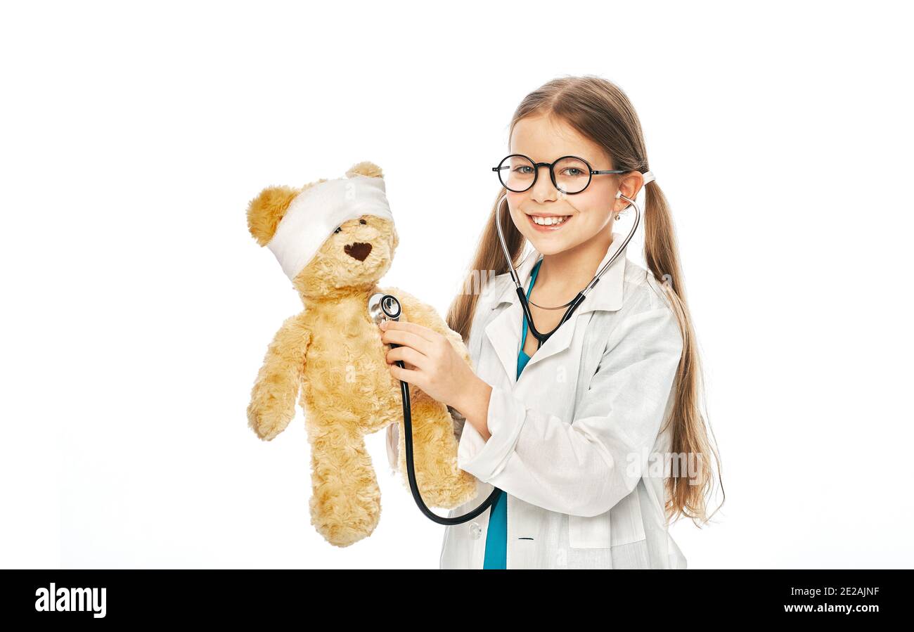 Caucasian girl wearing a doctor's coat plays with her toy bear in a medical game, using phonendoscope. White background Stock Photo