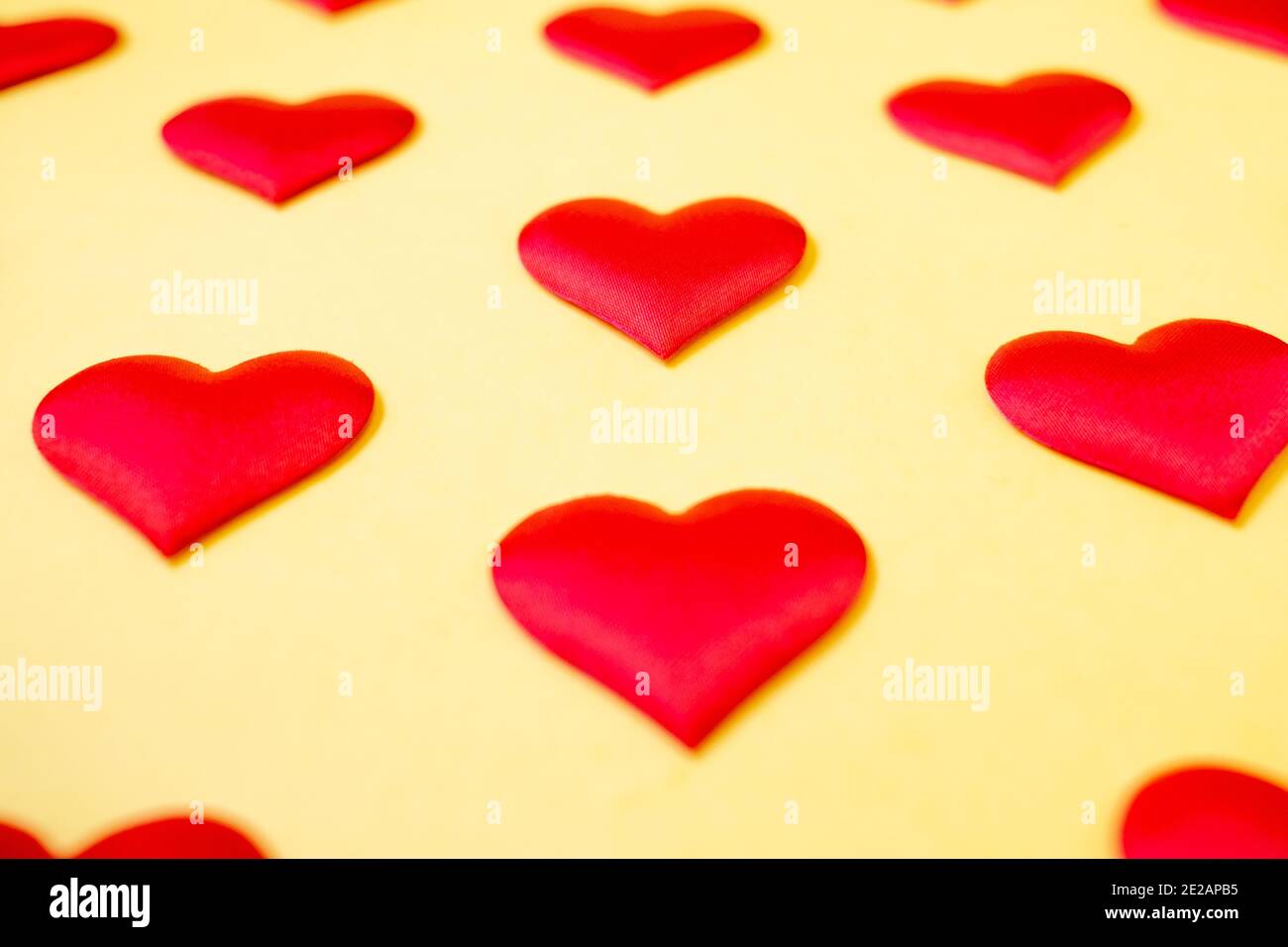 A lot of identical red silk hearts lying staggered on a yellow background. Symbol of love, tenderness and passion. Stock Photo