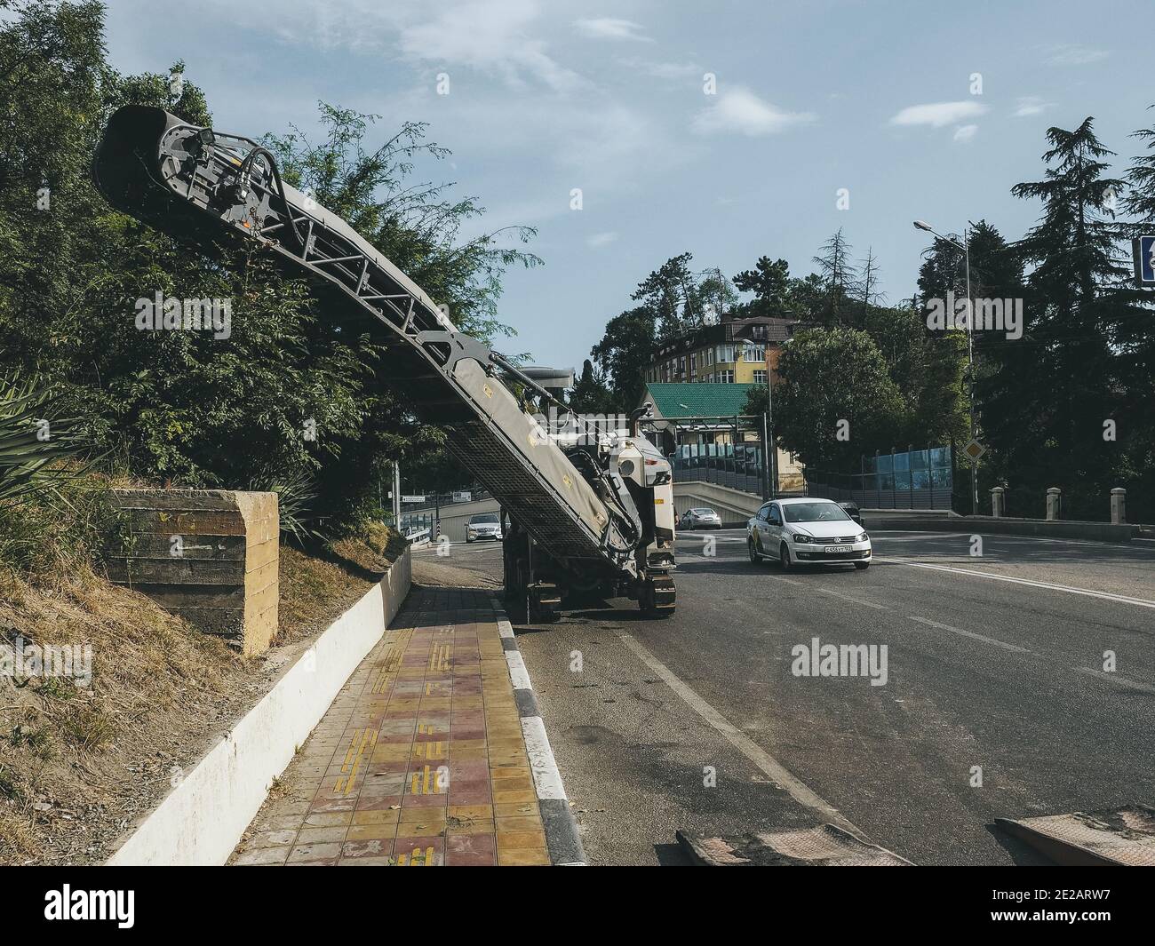 Russia, Sochi 05.07.2020. A road milling machine stands on the side of road in a city with passing cars and green trees in the background Stock Photo