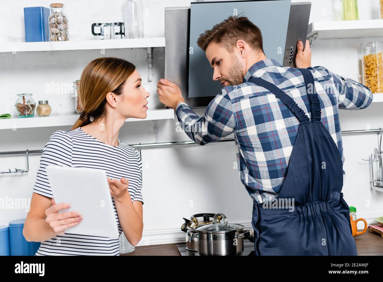 young handyman looking at tablet while repairing extractor fan near woman in kitchen Stock Photo