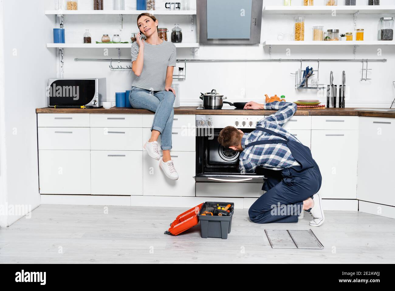 full length of smiling woman talking on smartphone while sitting on table near handyman repairing oven in kitchen Stock Photo