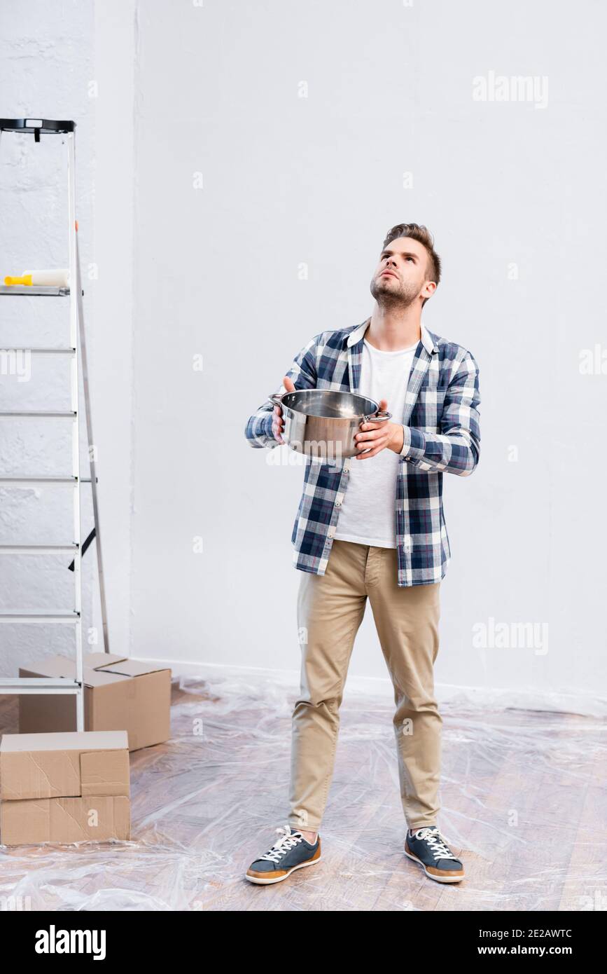 full length of young man with saucepan looking up while standing near cardboard boxes and ladder under leaking ceiling at home Stock Photo