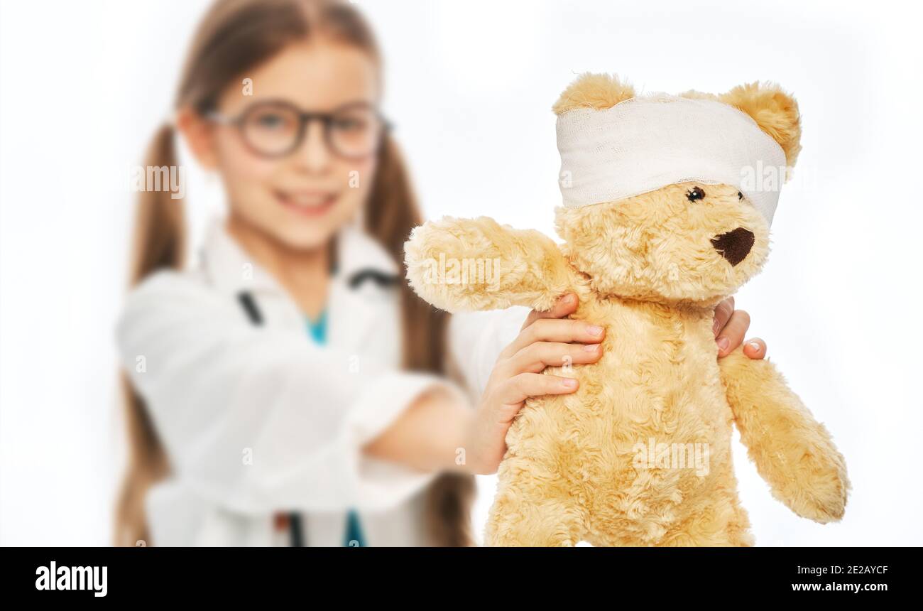 Little girl wearing a medical coat holding her toy bear with a bandaged head. Animal care and assistance, veterinarian Stock Photo