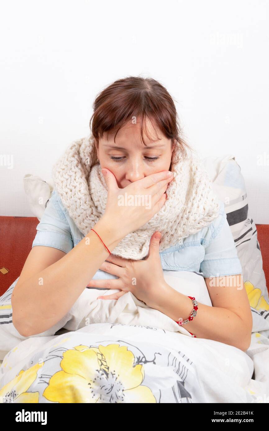 a young pretty girl is sick at home, lies in bed, coughs and holds on to her chest and covers her mouth with her hand Stock Photo