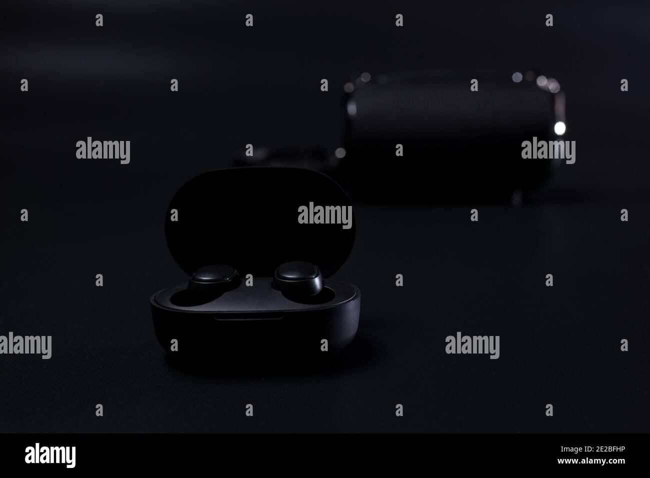 Black wireless headphones in charging case on black with wireless speaker on background. Stock Photo