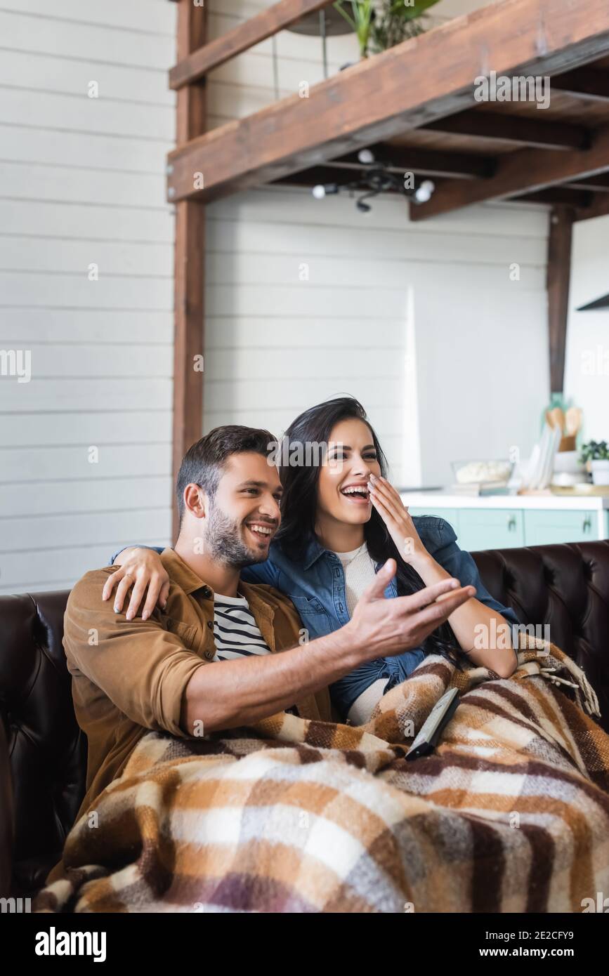 cheerful man pointing with hand near laughing woman covering mouth while watching tv under plaid blanket Stock Photo