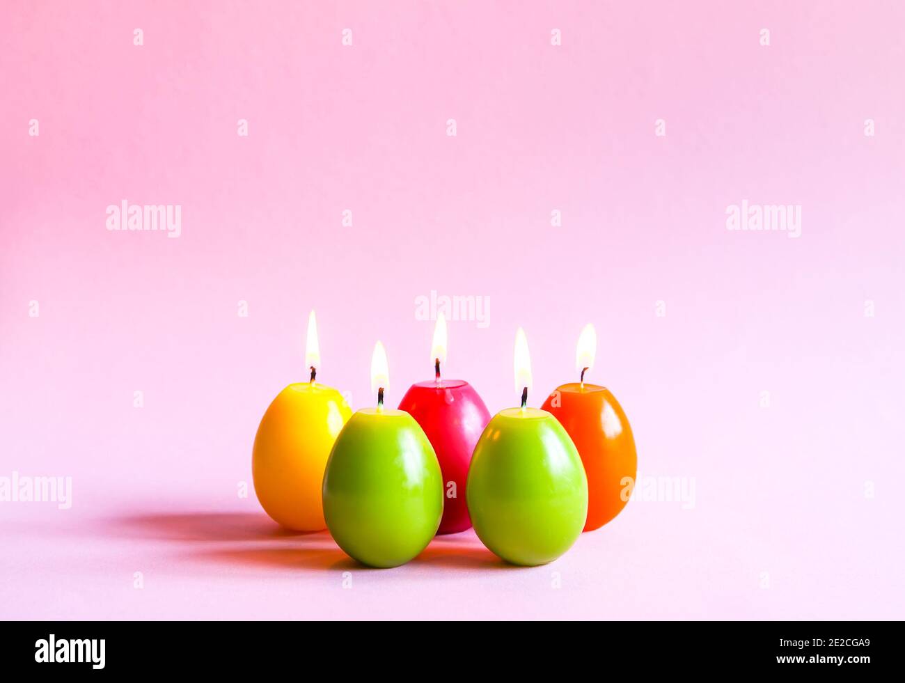Traditional Easter decor. Group of bright burning paraffin candles in the shape of colorful eggs on soft pink background. Stock Photo