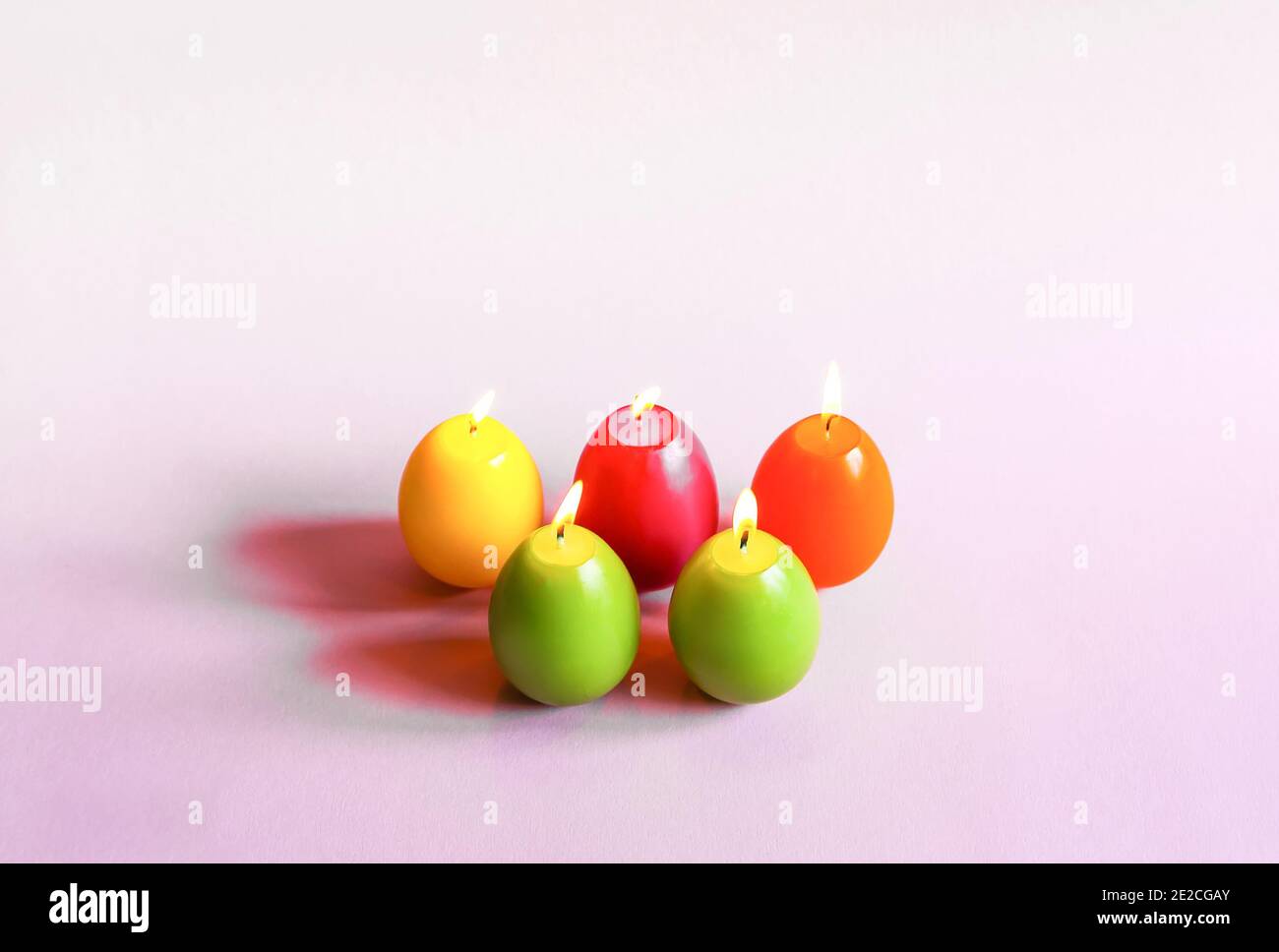 Traditional Easter decor. Group of bright burning paraffin candles in the shape of colorful eggs on soft pink background. Stock Photo