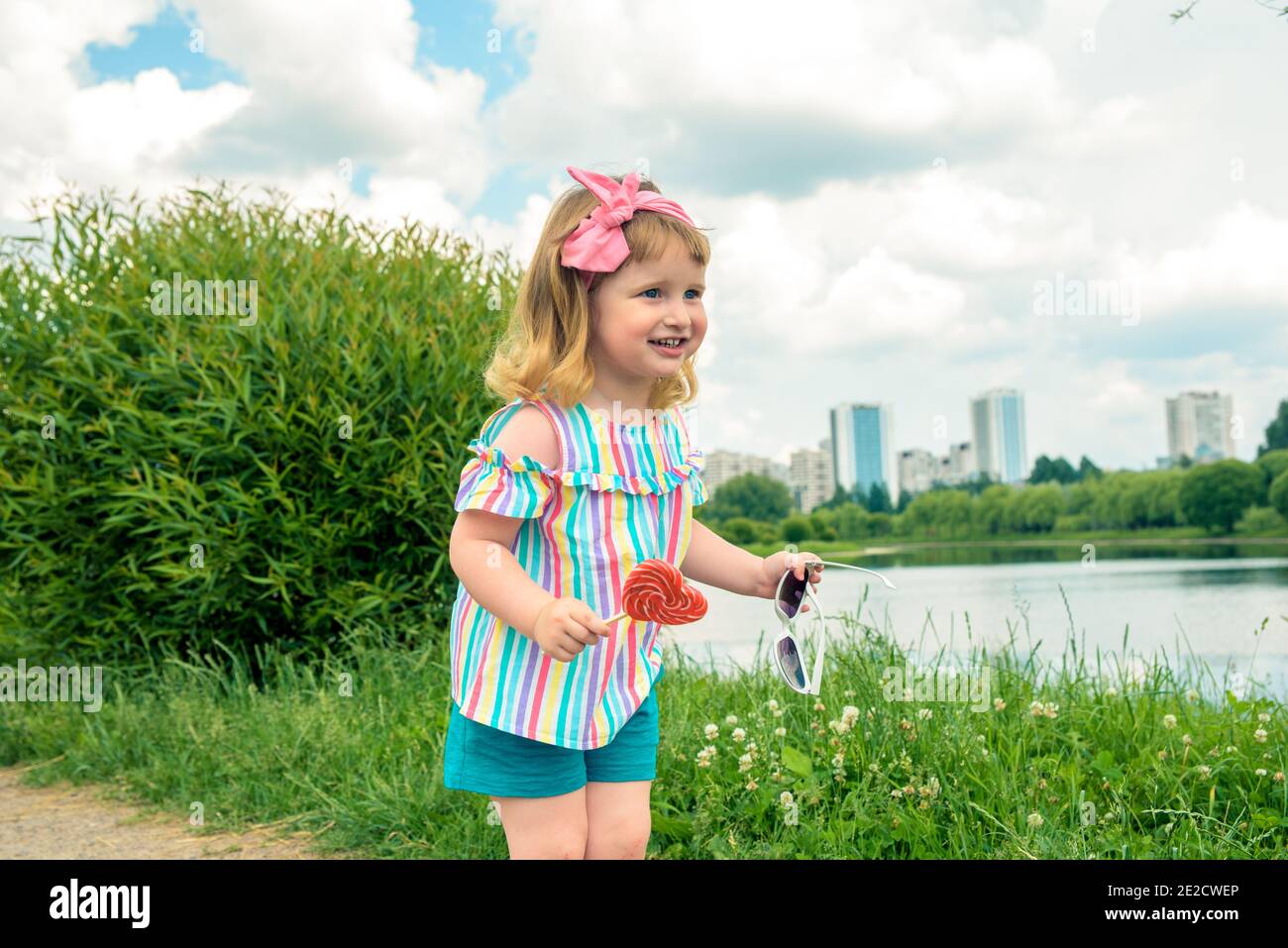 Cute curly baby girl eating watermelon candy in a sunny park. baby with outdoors lollipop Stock Photo