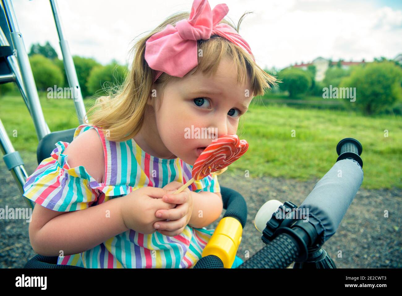 Cute curly baby girl eating watermelon candy in a sunny park. baby with outdoors lollipop Stock Photo