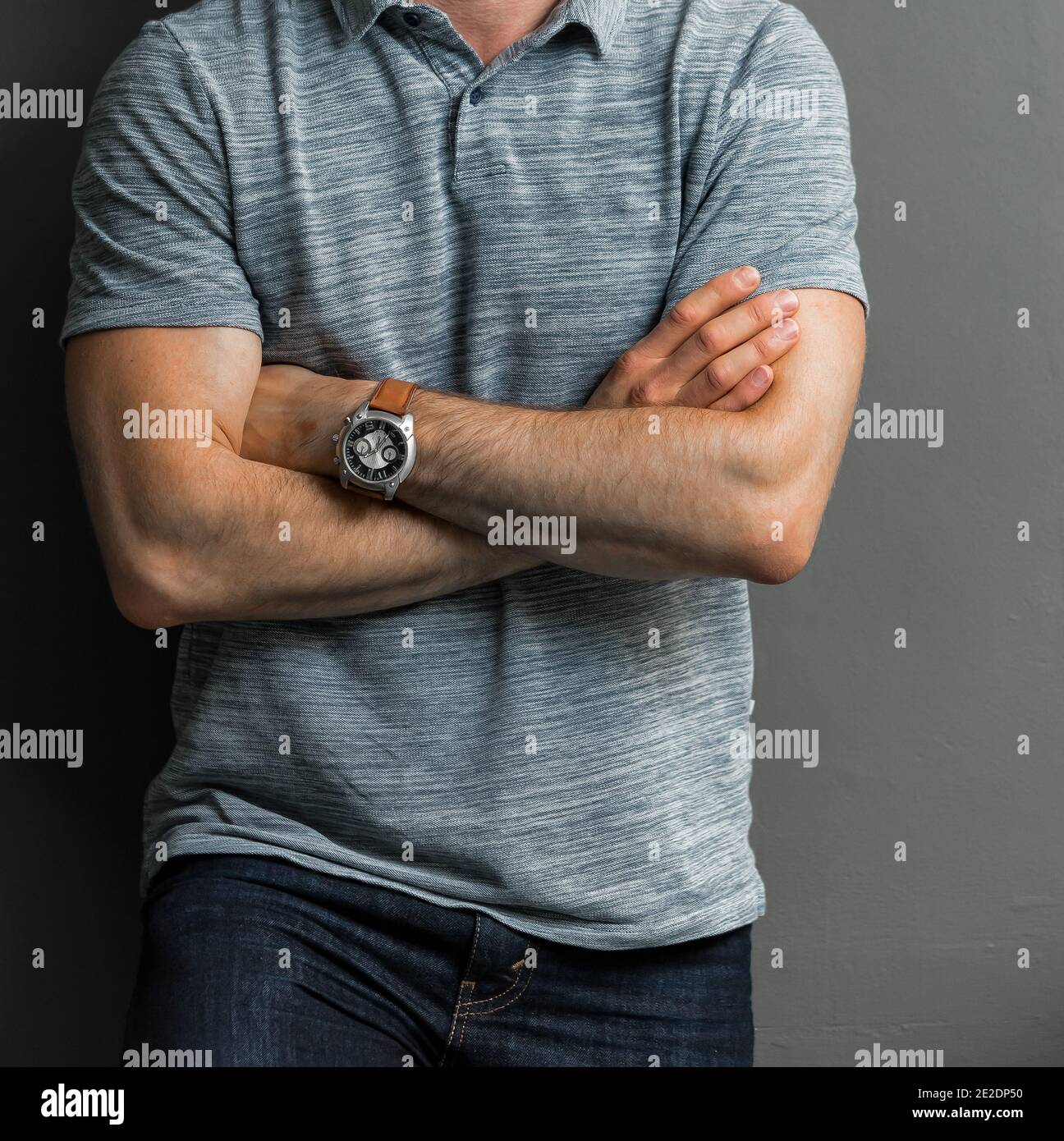 Young man in a gray shirt wearing a watch with his arms cross leaning on a wall Stock Photo