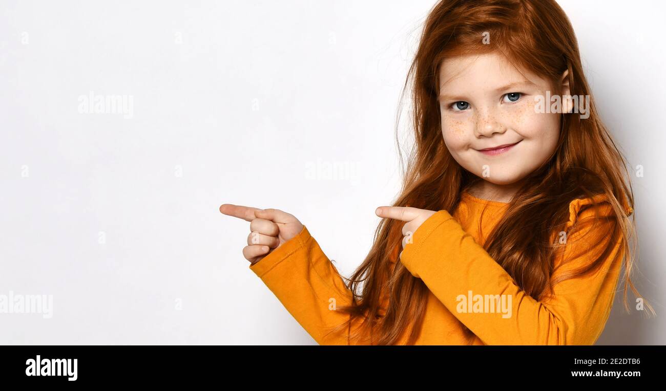 Smiling small red haired girl with freckles in yellow comfortable longsleeve standing and pointing aside with fingers Stock Photo
