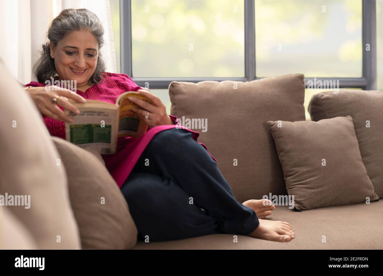 AN OLD WOMAN HAPPILY READING A BOOK WHILE SITTING COMFORTABLY ON SOFA Stock Photo