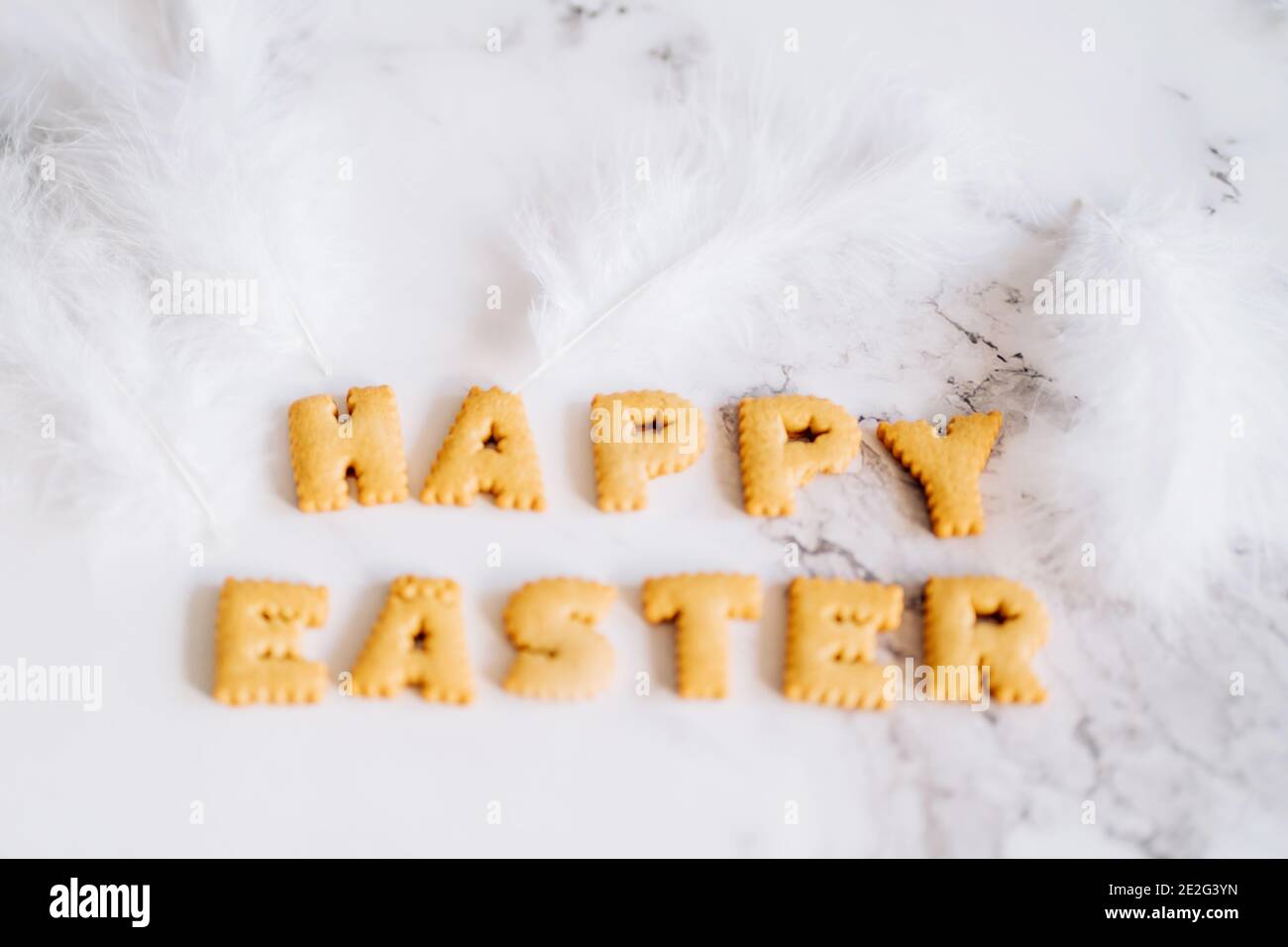 Lettering from biscuits in form of letters and white fluffy plumelets on marble background. Happy Easter background, creative greeting. Stock Photo